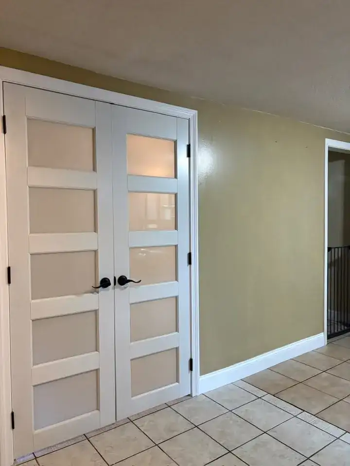 A pair of new white French doors with five frosted glass panels each, black lever handles and fresh casing, on a beige tile floor.