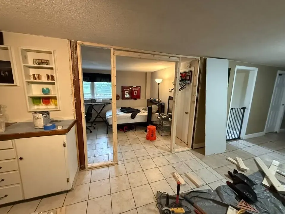 A new rough opening framed in raw lumber in a finished basement, with the bedroom visible through it and tools on the floor.