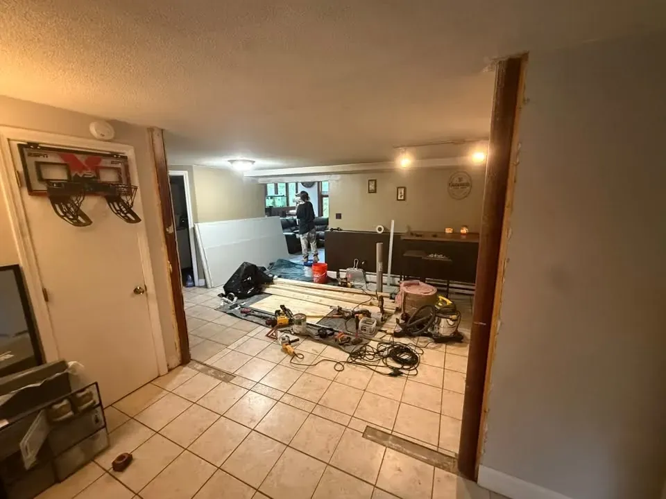 A wide opening in a basement wall with a wood header, and a room beyond full of tools, cords and materials on a tile floor.