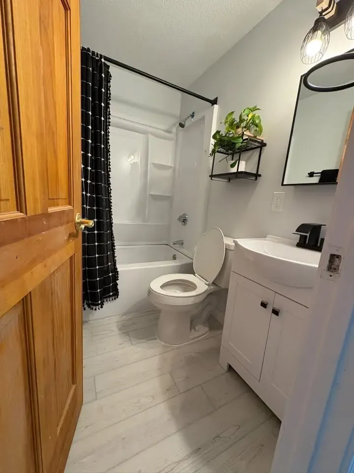 A finished small bathroom with a white tub surround, plaid shower curtain, white vanity, round black mirror and light grey plank floor.