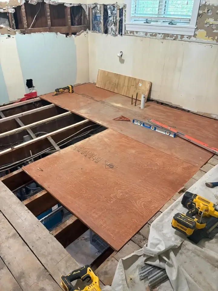 A floor opened down to the joists, with new plywood subfloor sheets going down and a cordless drill on the boards.