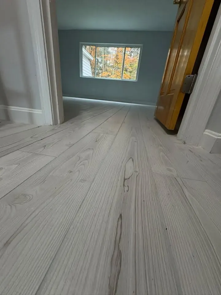 A bedroom with new light grey wide-plank flooring running toward a window with autumn trees outside.
