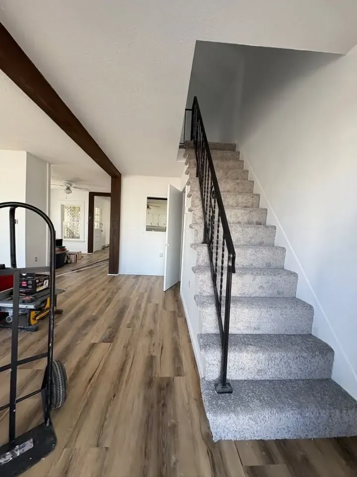 An entry hall with warm grey-brown plank flooring, a carpeted staircase with a black iron railing and a dark beam overhead.