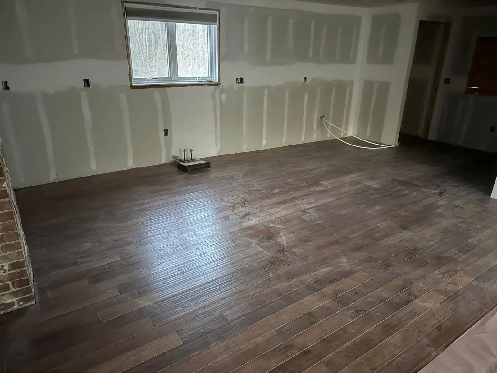 A room with new dark brown plank flooring and freshly taped drywall on the walls around a small window.