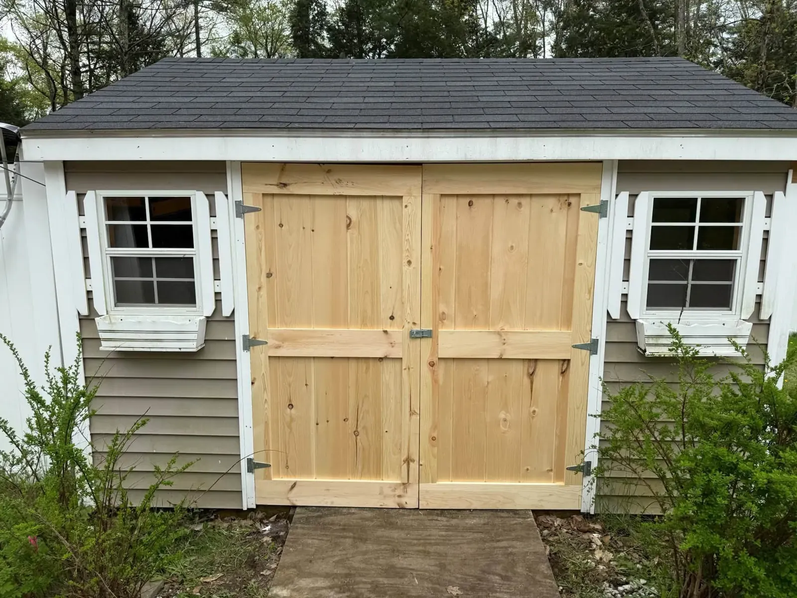 A grey-sided shed with new double doors built from natural pine boards, white trim and two small white windows.