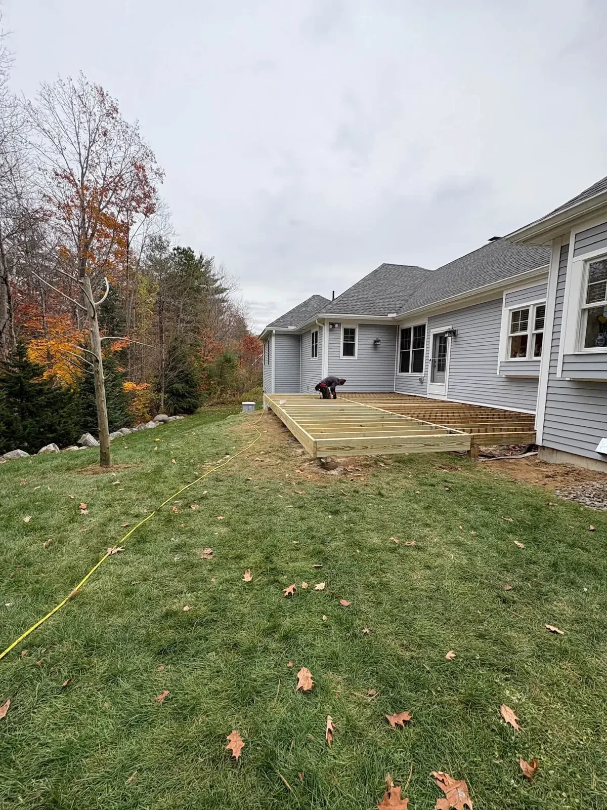 A low deck frame on a lawn behind a grey-sided ranch house, with a string line staked out and fall trees behind.