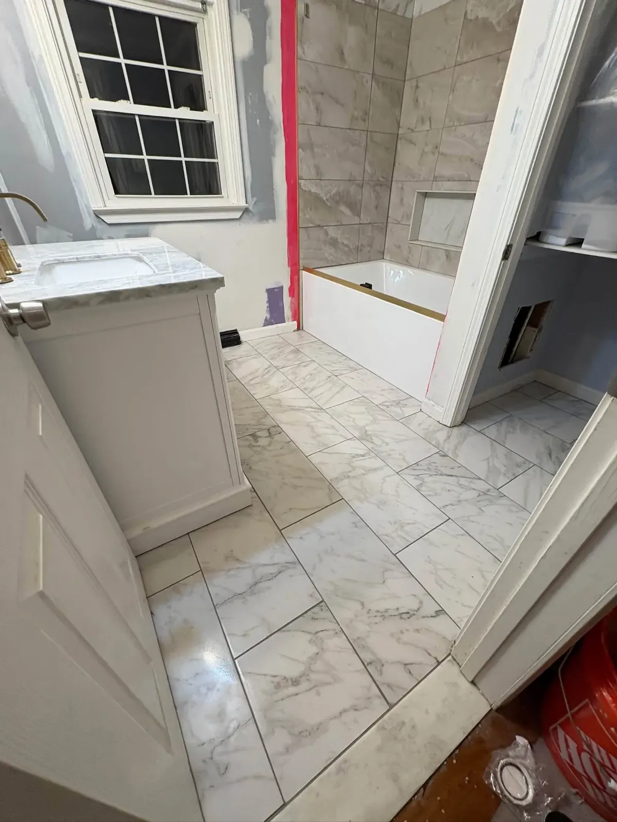 A bathroom with a new large-format marble-look tile floor, a grey tile tub surround going in and a white vanity.