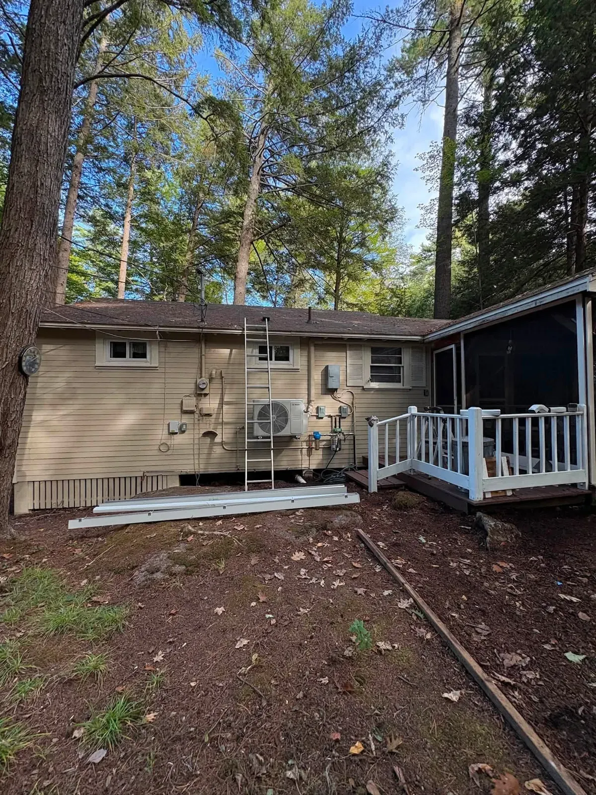 The back of a small cottage with beige vinyl siding, a screened porch and a white-railed deck among tall pines.