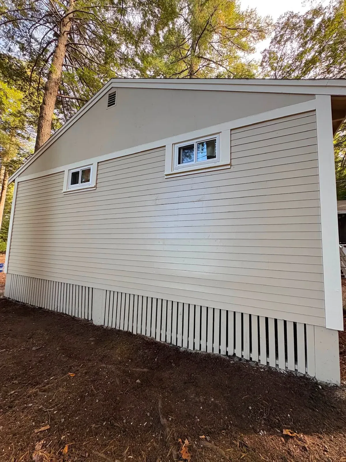 The gable end of a cottage with new beige vinyl siding, white trim, two small windows and white lattice skirting.