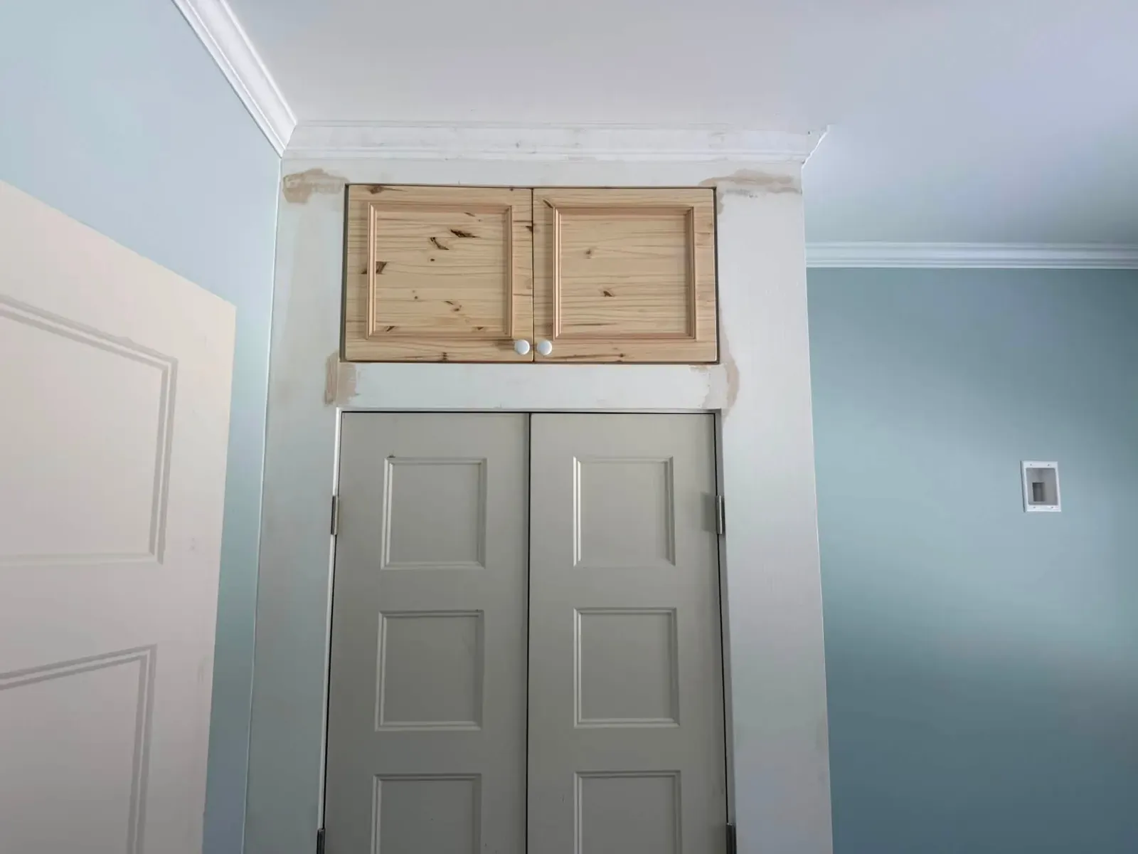 A closet with new grey panel doors under a small pine cabinet, framed in white casing and crown on a pale blue wall.