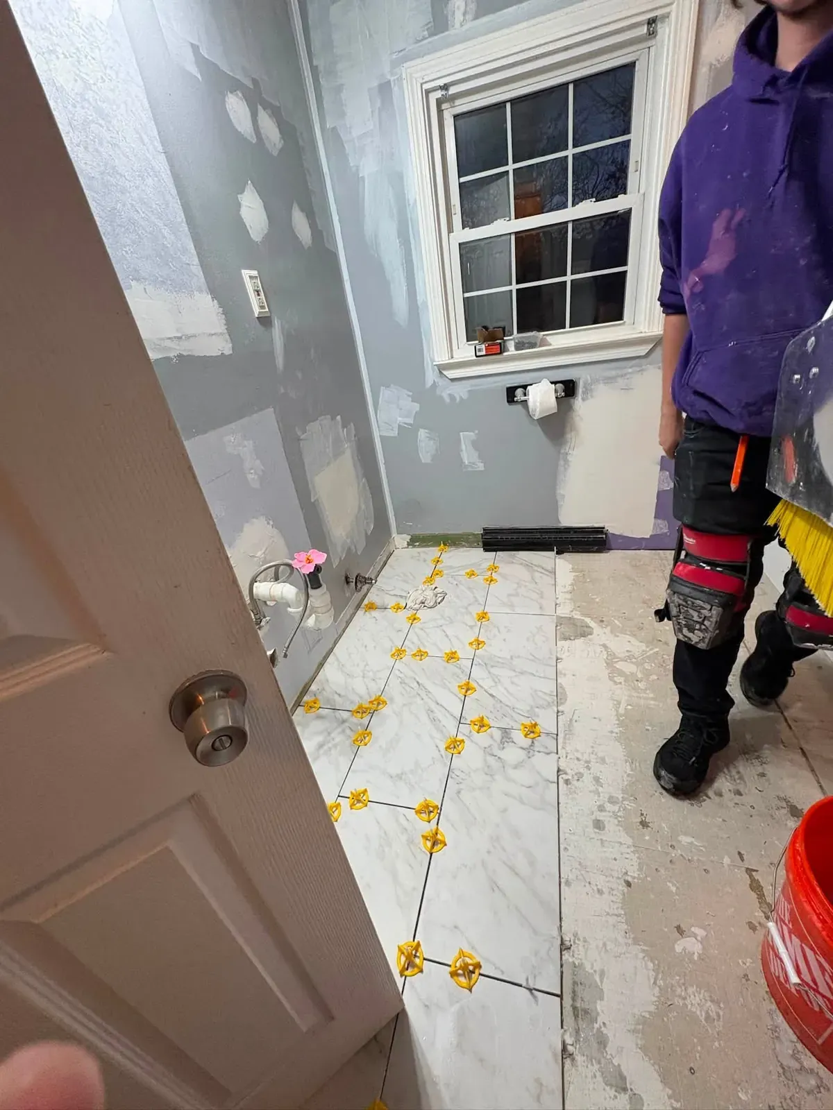 Marble-look floor tile being laid in a bathroom with yellow leveling clips at every joint and a worker standing in the doorway.