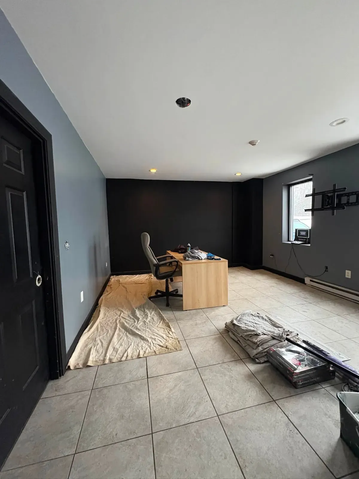 A room painted blue with one charcoal accent wall, a large tile floor and a desk set up under the window.