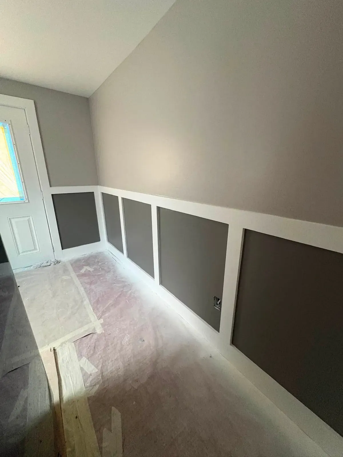 A hallway with new board-and-batten wainscoting, white rails over charcoal panels, above a paper-covered floor.