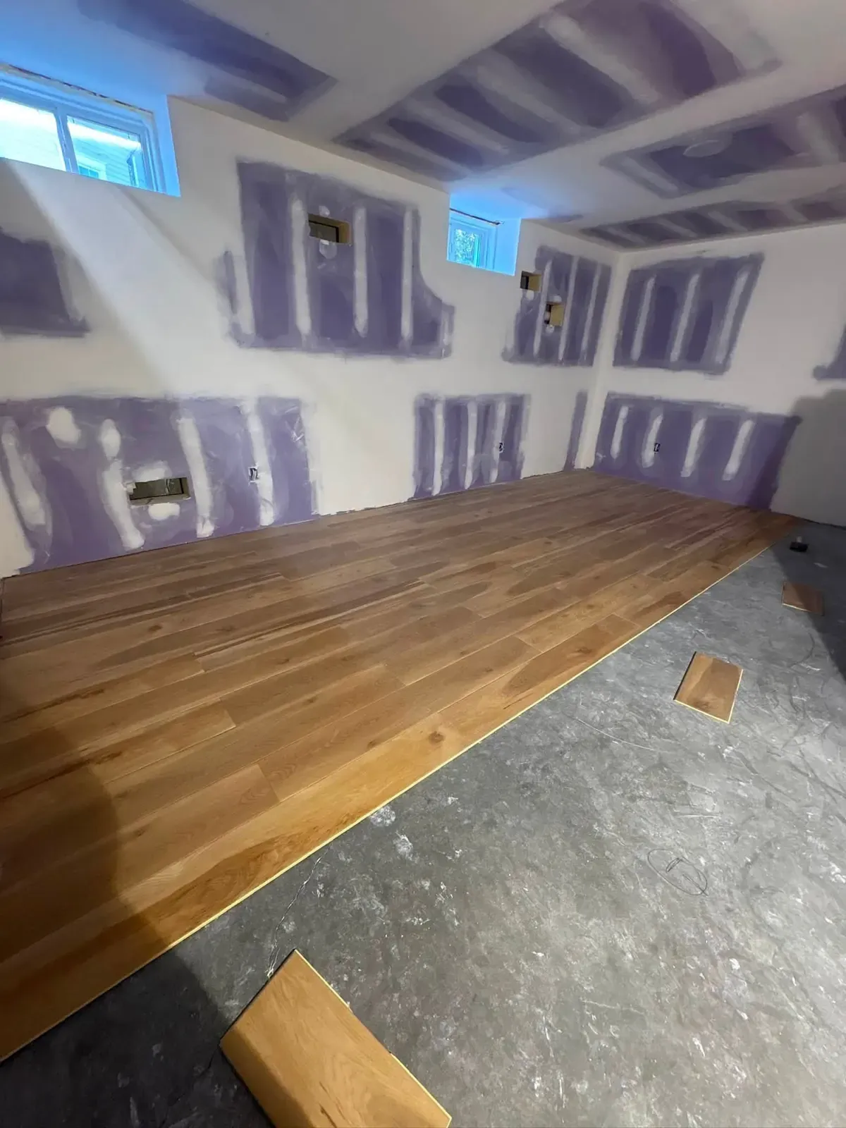 A basement room with new purple drywall on the walls and ceiling, screws mudded, and oak plank flooring half laid over the slab.