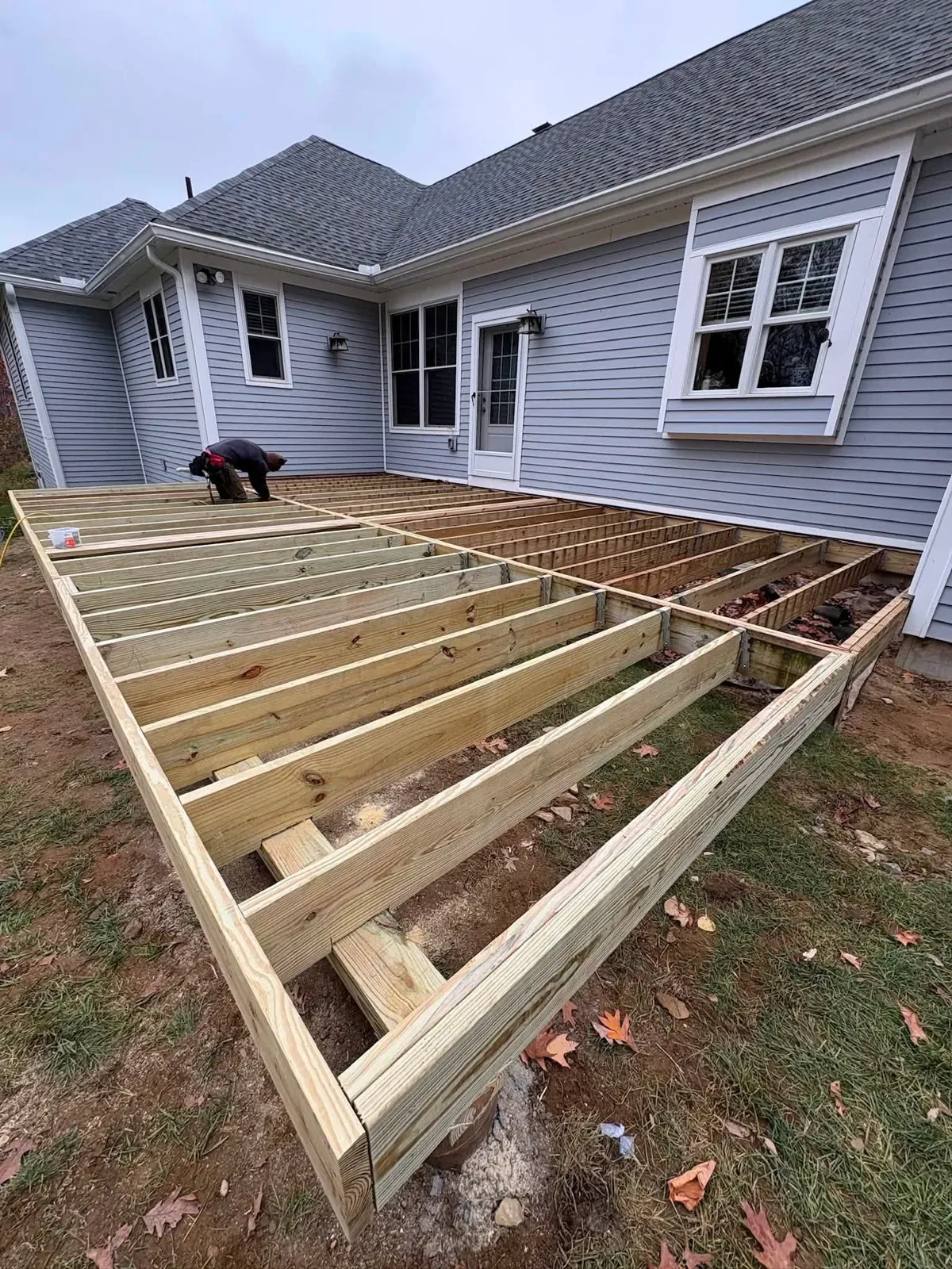 A large pressure-treated deck frame with joists set on a lawn behind a blue-sided house, with a worker kneeling at the far end.