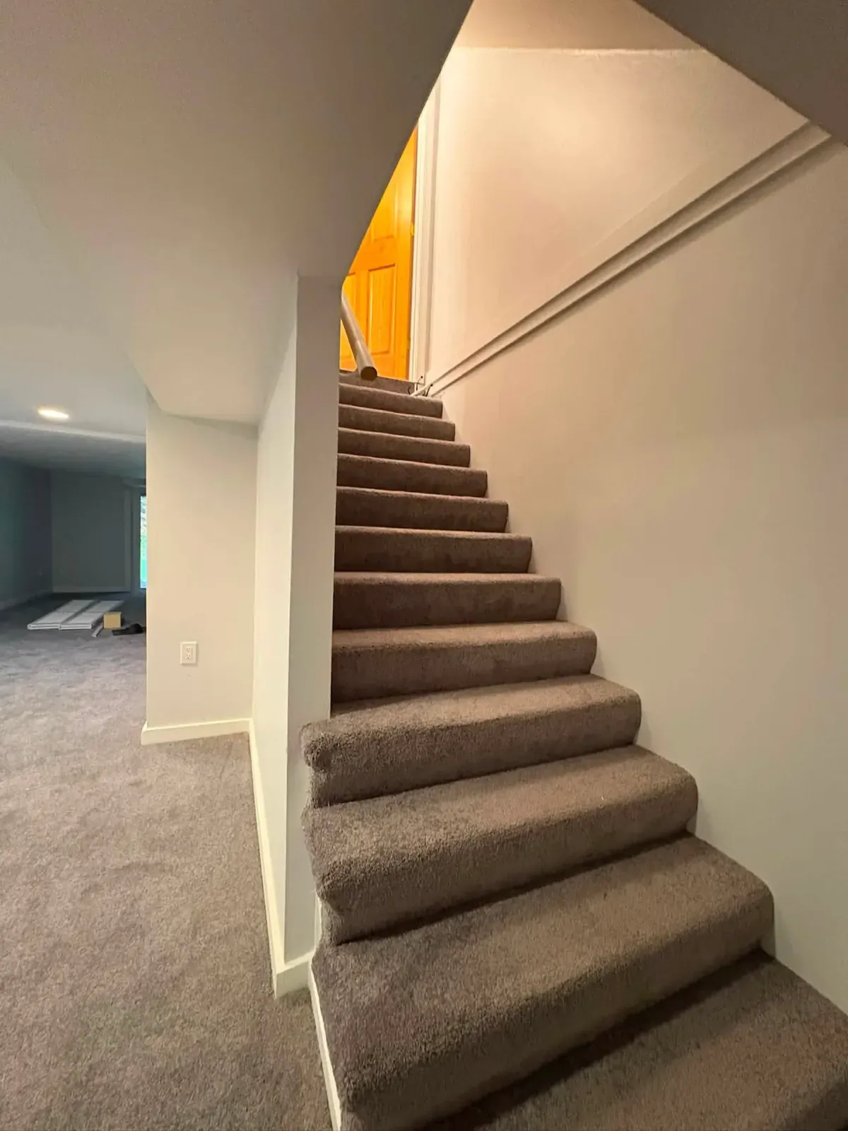 A carpeted staircase rising from a finished basement with white walls, white trim and light carpet.